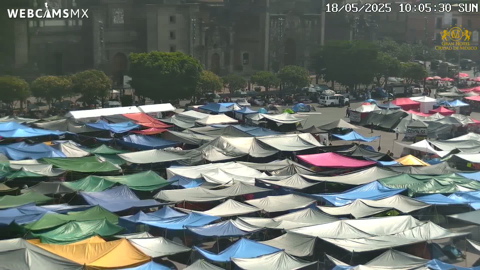 A large number of tents of various colors are densely packed together in a sunny outdoor plaza in front of a large building.