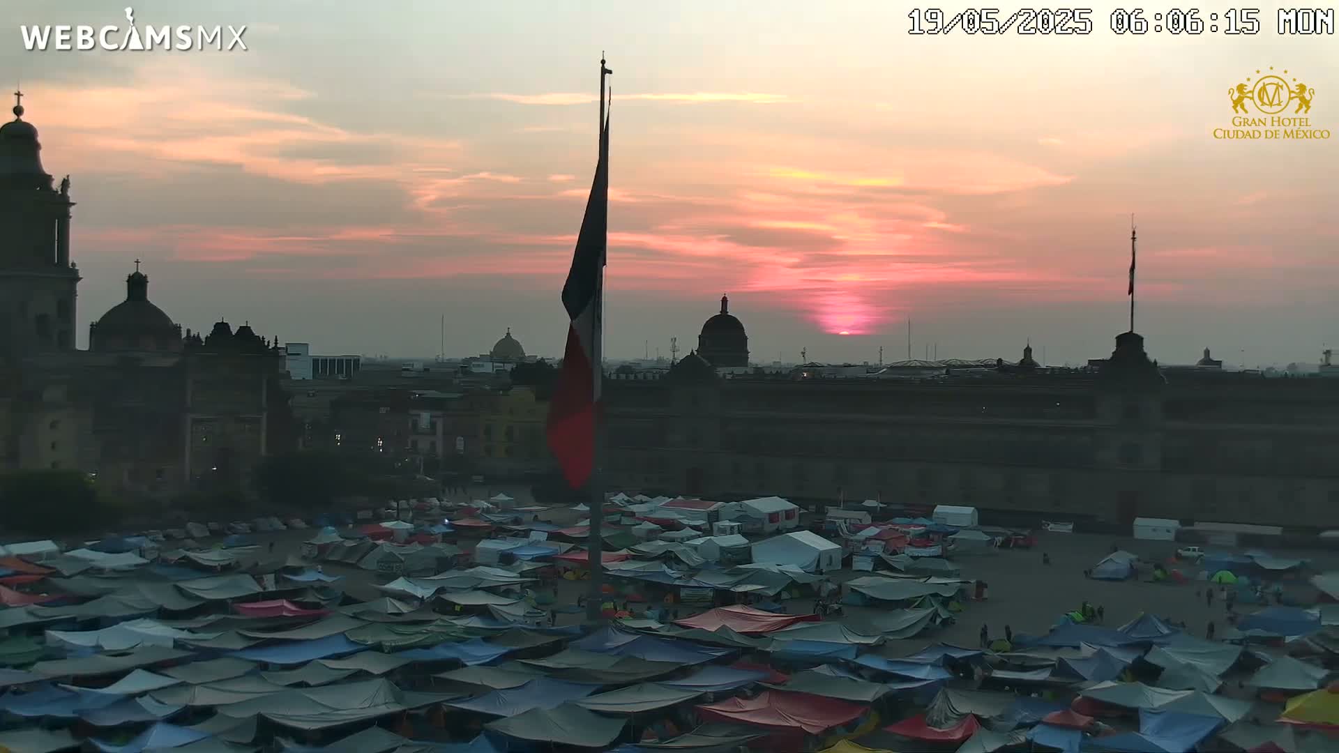A large plaza filled with numerous tents is seen at sunset, with a prominent building and flagpole in the background.