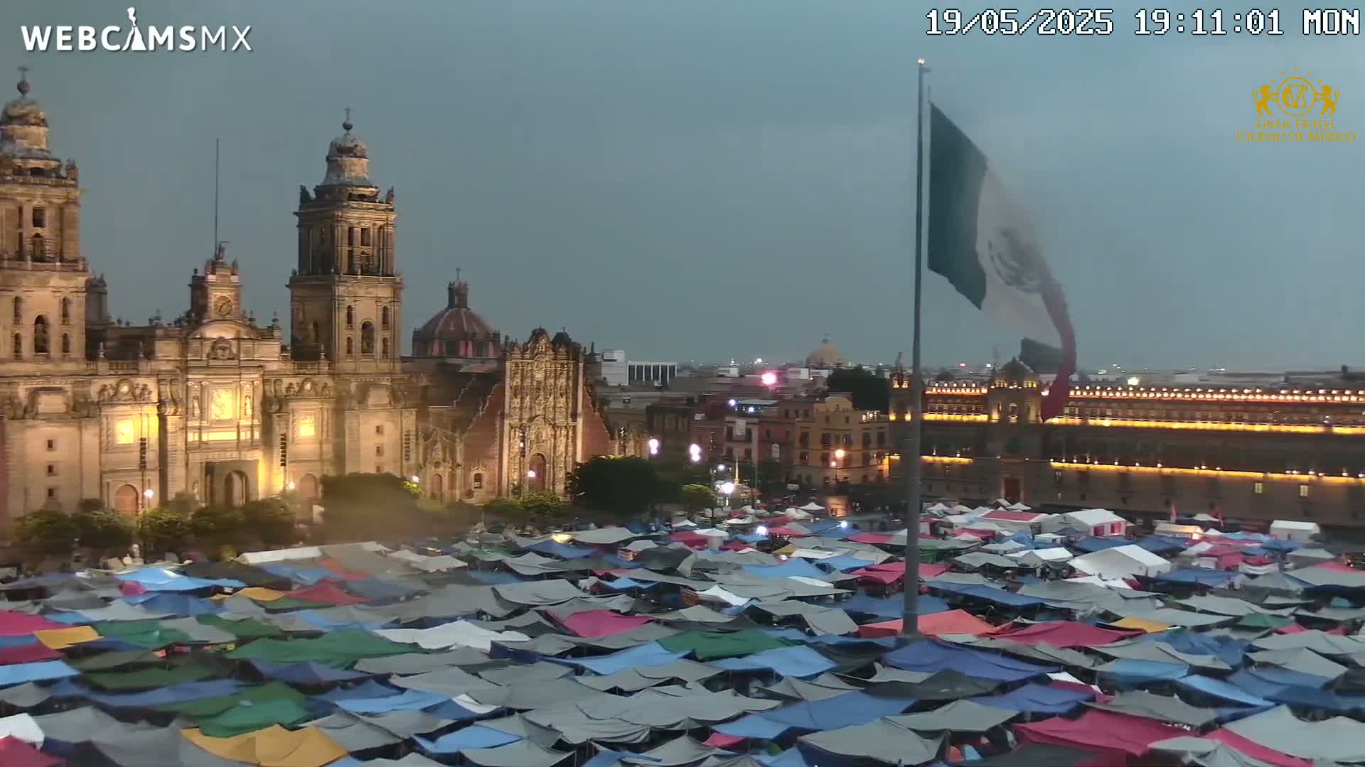 A large plaza at twilight, filled with numerous tents in front of a brightly lit cathedral and government building under an overcast sky.
