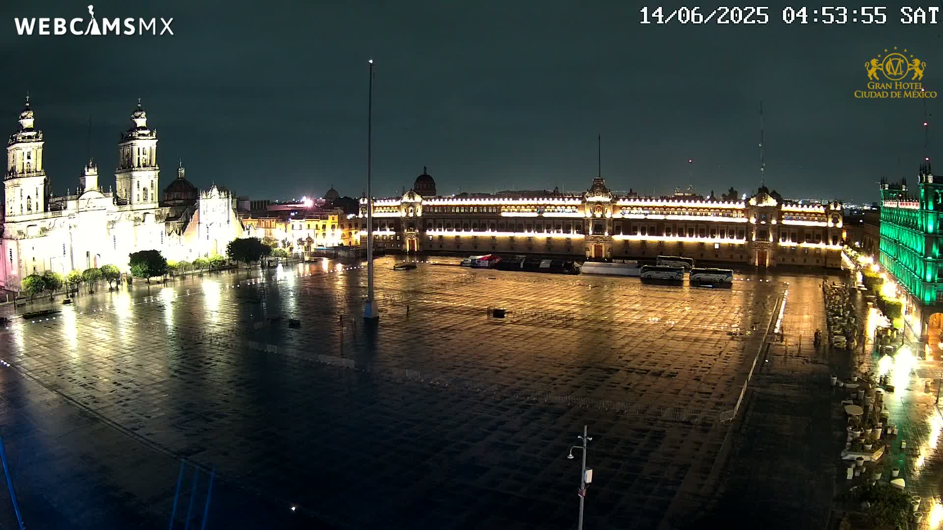A large, empty plaza at night is illuminated, showing a cathedral and government buildings in the background under a rainy sky.