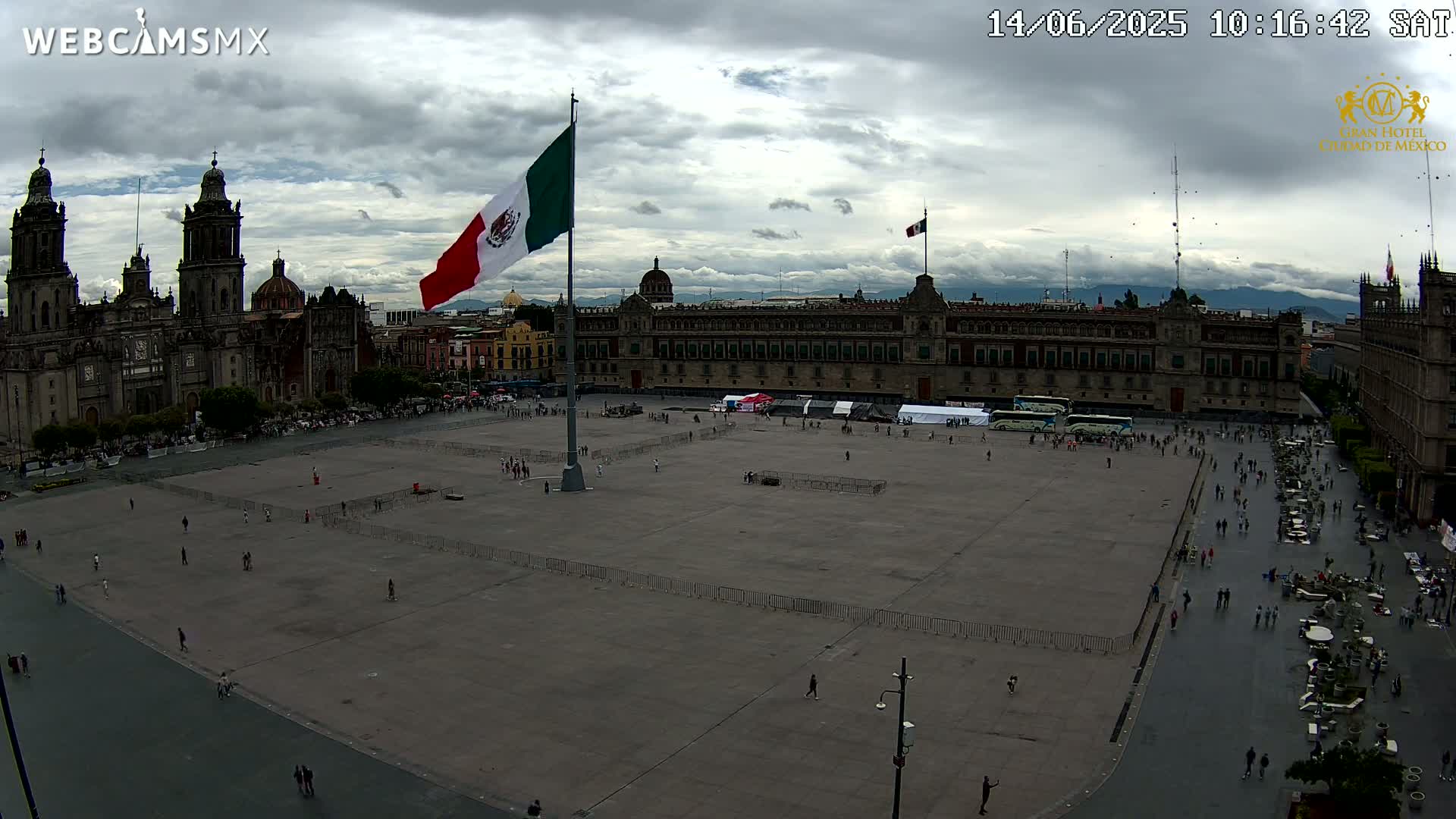 A large plaza, mostly empty except for scattered people, with a large Mexican flag on a tall pole and a cathedral in the background under a cloudy sky.