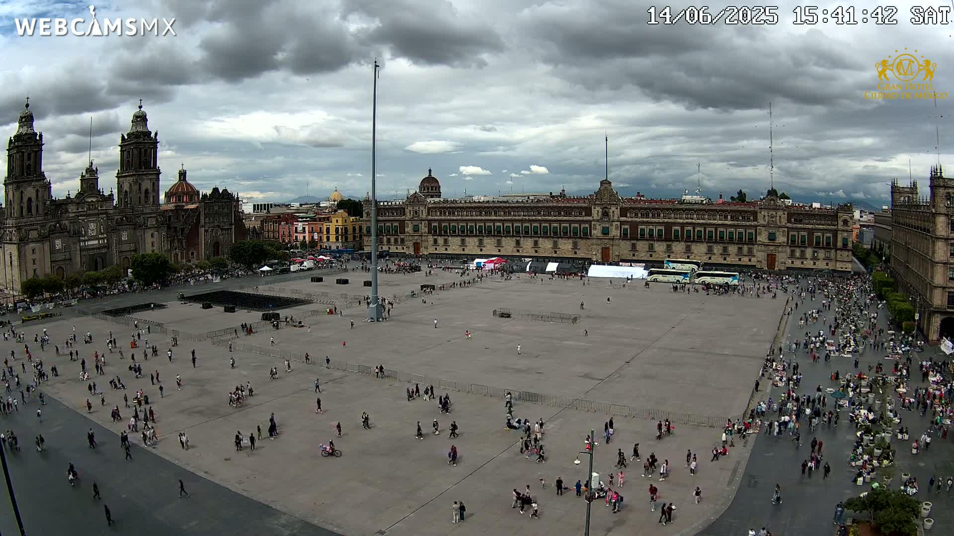 A large plaza, mostly empty except for scattered people, with a large Mexican flag on a tall pole and a cathedral in the background under a cloudy sky.