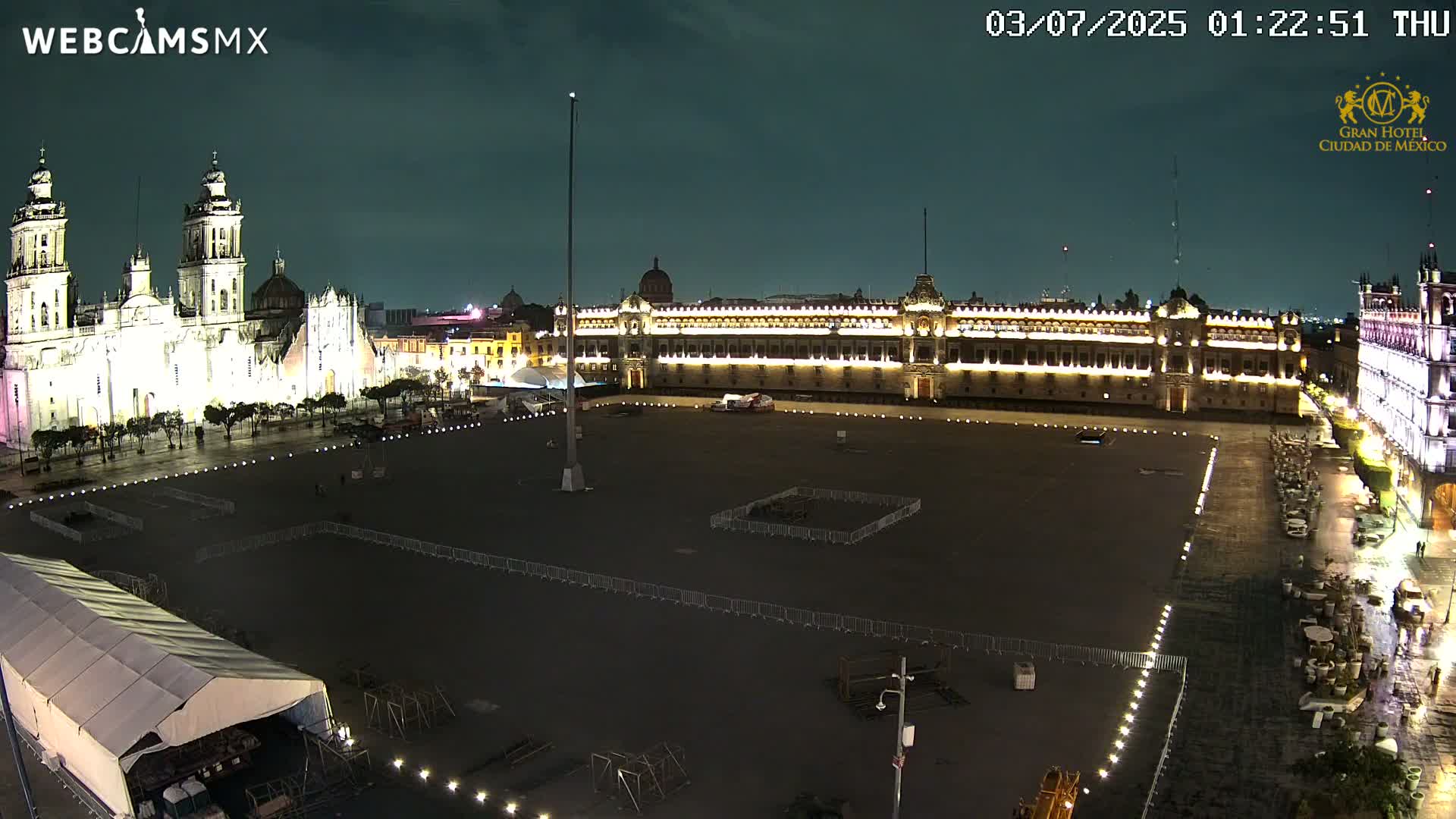 A large, mostly empty plaza at night, illuminated by lights, is bordered by a cathedral and government buildings.