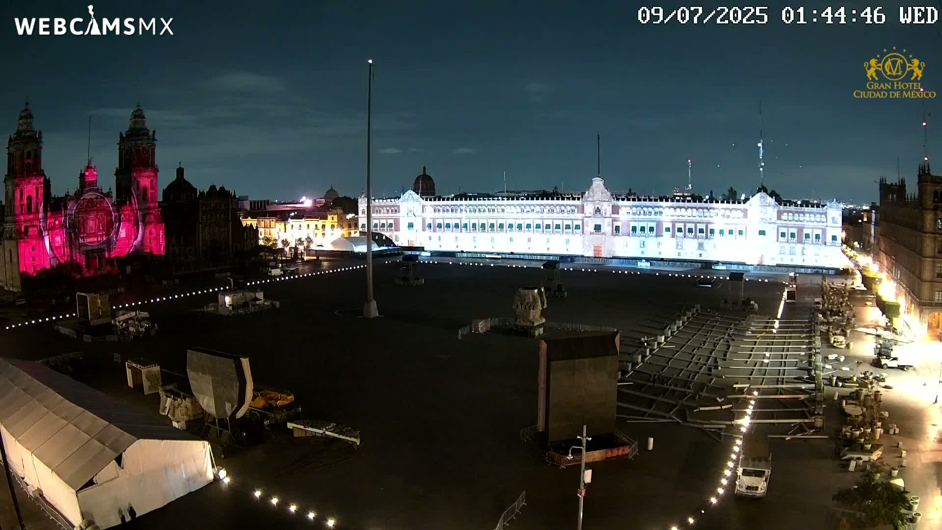 A large plaza at night, illuminated by lights on buildings and the ground, shows a cathedral with pink projections, a large palace, and preparations underway for an event.