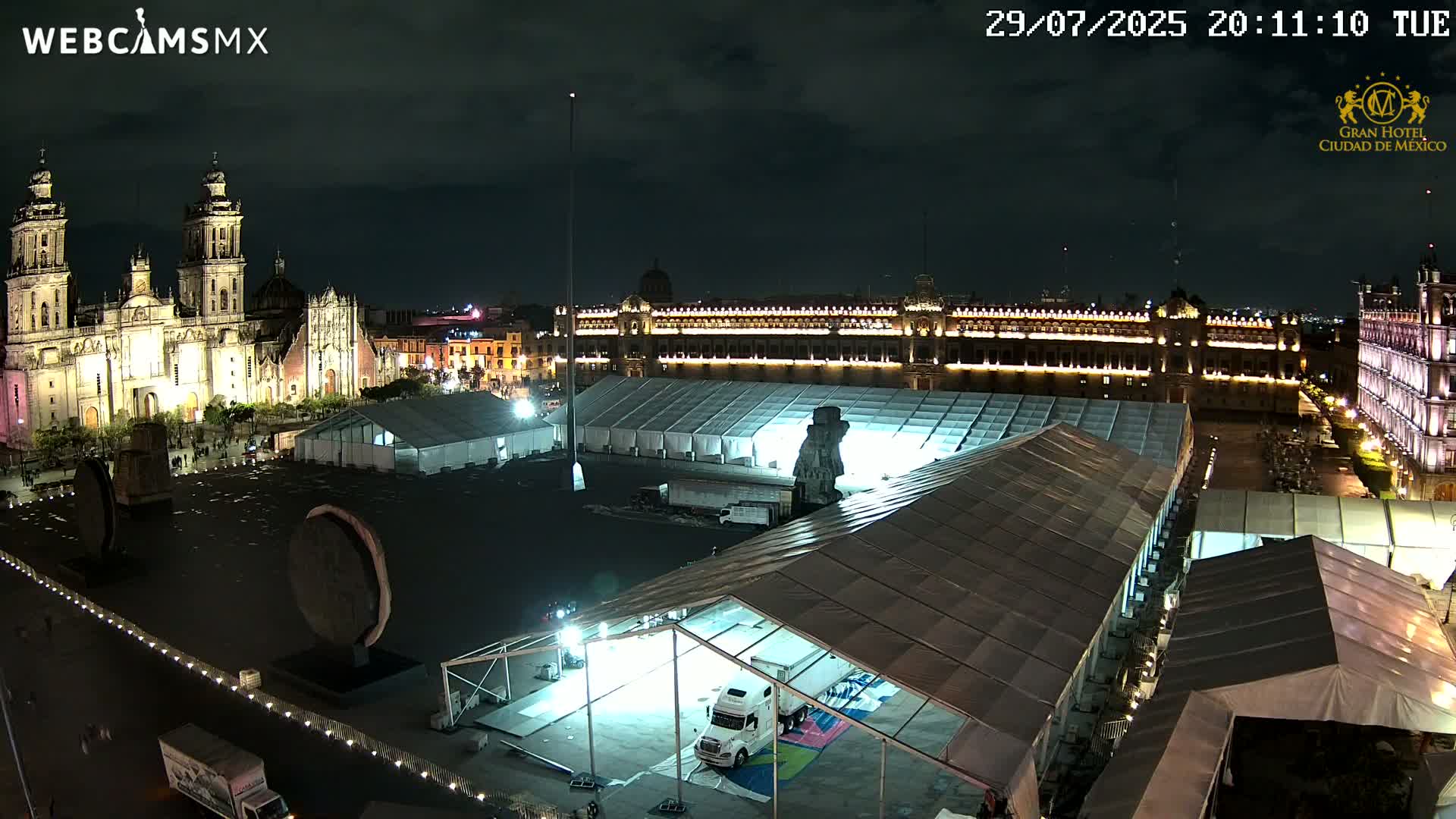A large plaza at night, illuminated by lights on buildings and the ground, shows a cathedral with pink projections, a large palace, and preparations underway for an event.
