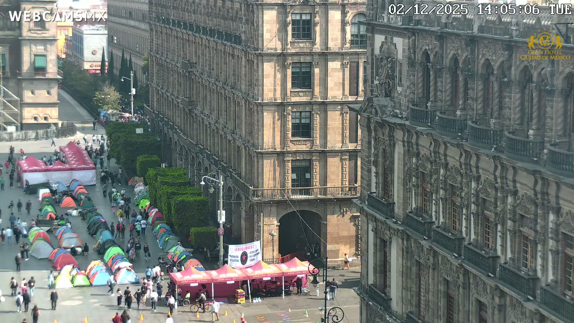 A bustling urban plaza is filled with numerous people walking among rows of colorful camping tents and larger pink canopies, all set against a backdrop of grand, historic multi-story buildings under a bright, clear sky.