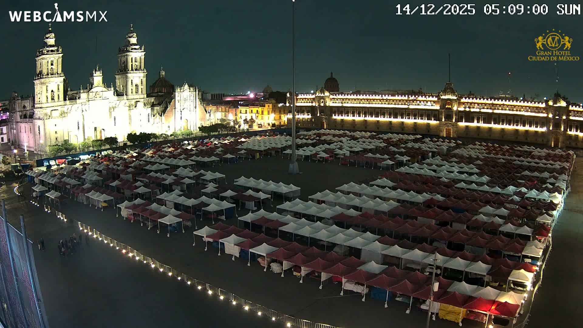 A bustling urban plaza is filled with numerous people walking among rows of colorful camping tents and larger pink canopies, all set against a backdrop of grand, historic multi-story buildings under a bright, clear sky.