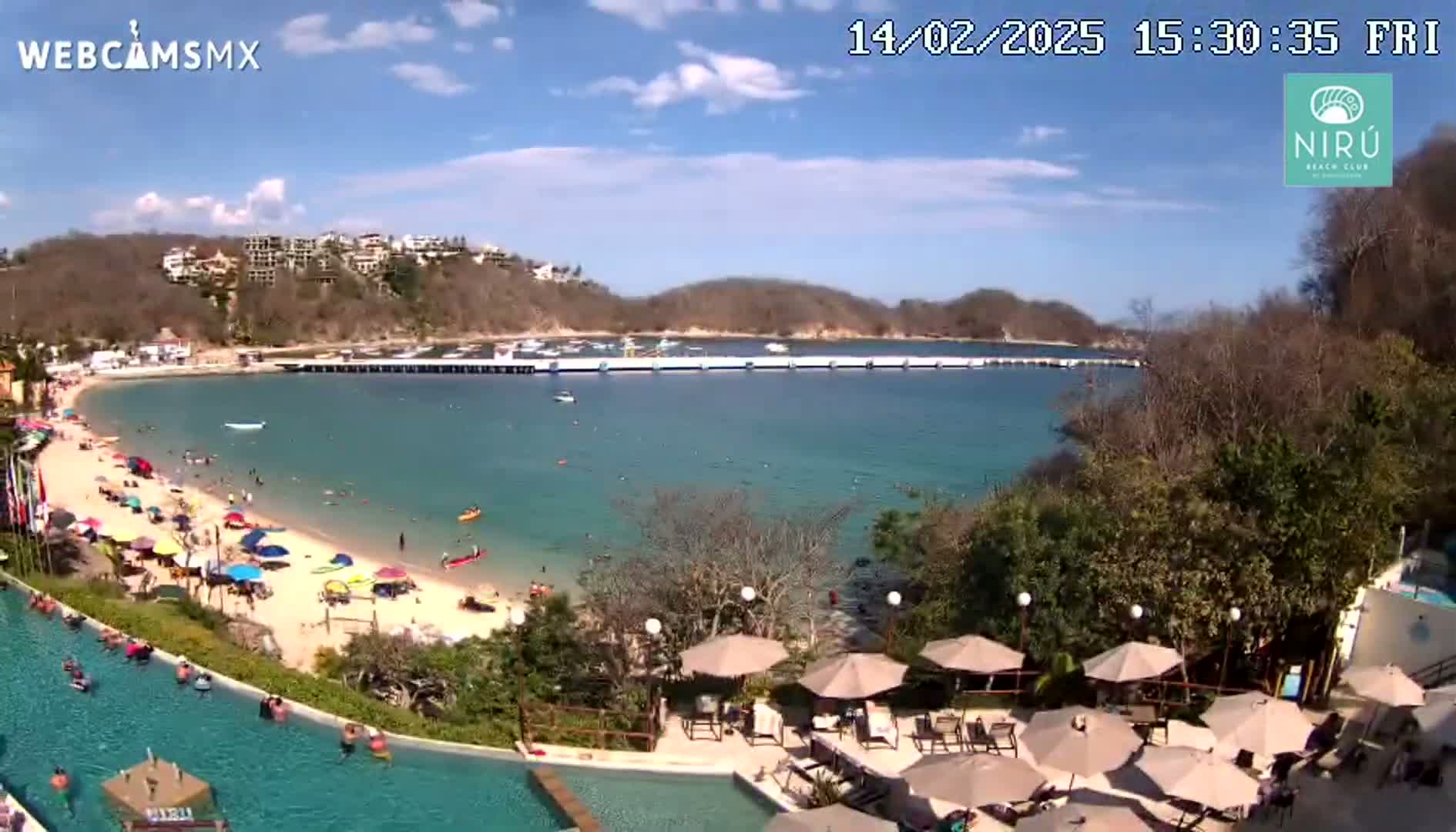 A sunny day at a beach resort shows a calm bay with a long pier, a sandy beach with people enjoying the water and sun, and a resort pool area with umbrellas and lounge chairs.