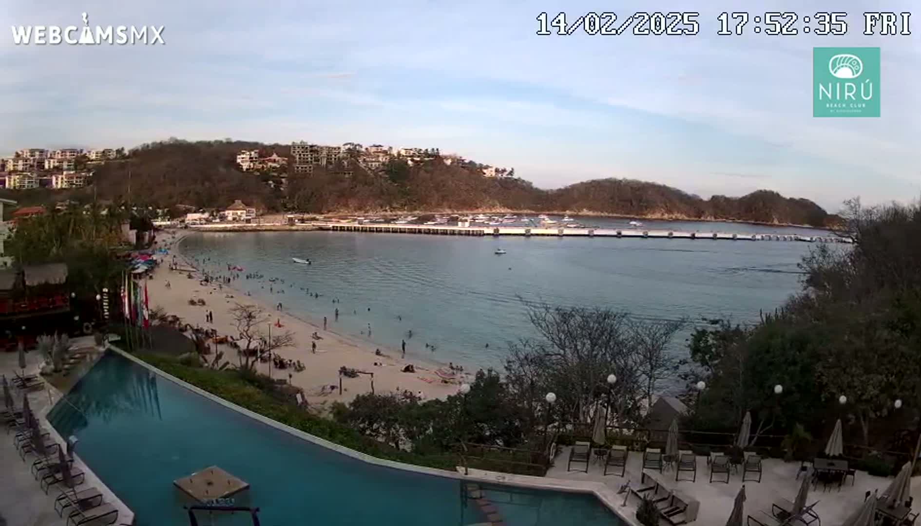 An aerial view shows a calm bay with a sandy beach crowded with people, a long dock, buildings on a hillside, and a resort pool area.