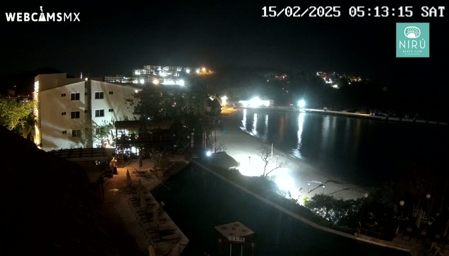 A nighttime view of a beachside resort with a swimming pool, illuminated buildings, and a sandy beach next to calm water.