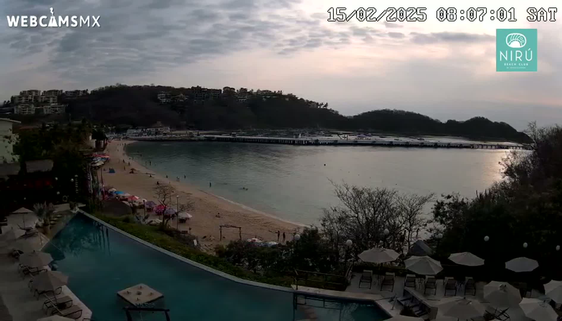An overcast evening view of a calm bay with a sandy beach, a long pier, buildings on a hillside, and a resort pool area with lounge chairs and umbrellas.