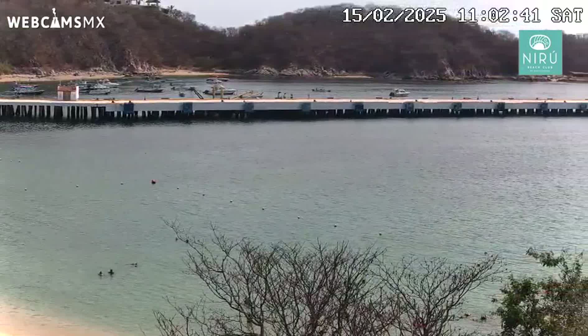 A long pier extends into a calm body of water, under an overcast sky, with several small boats moored nearby and a few people swimming in the bay.