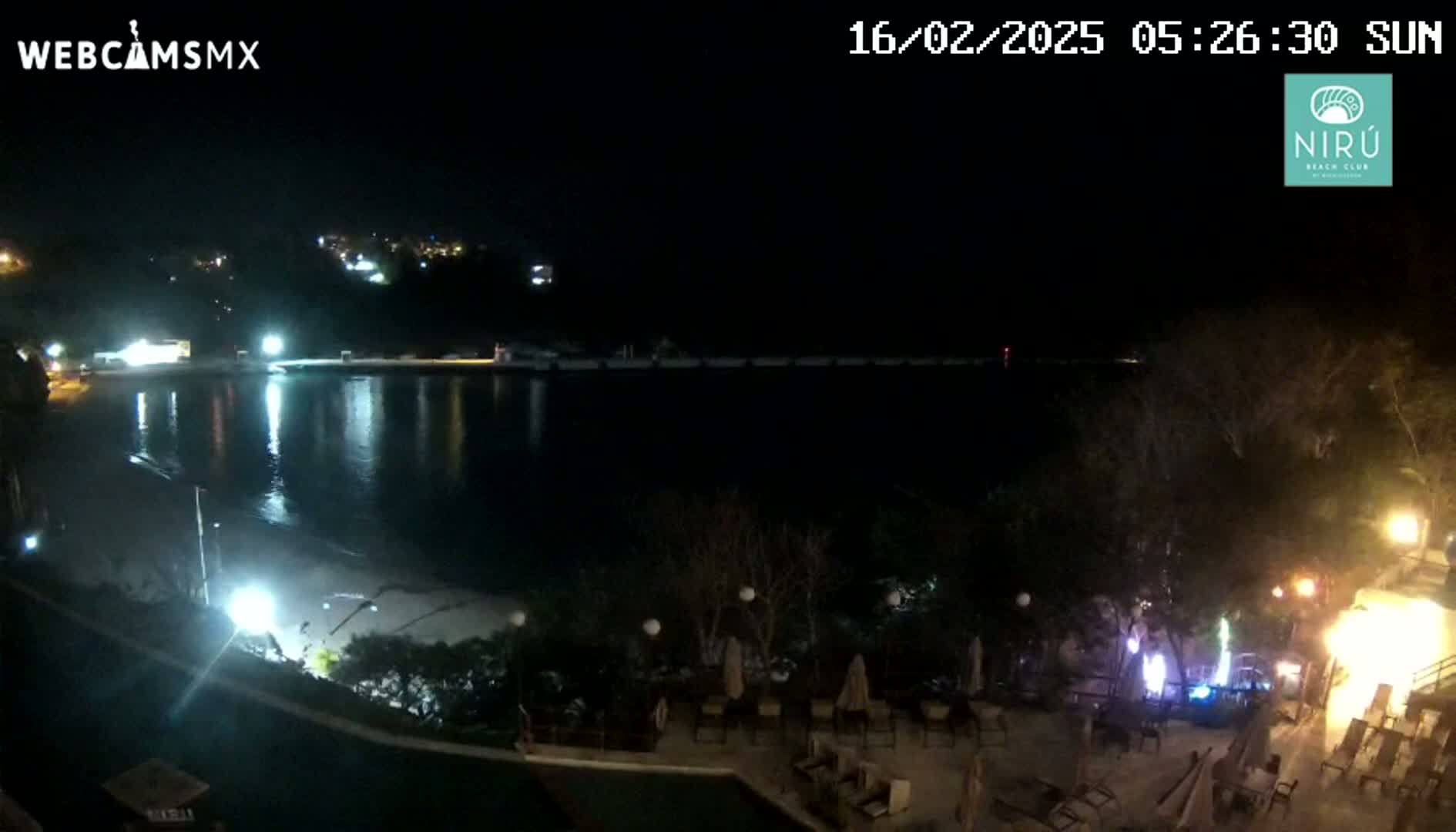 A nighttime view of a calm body of water reflecting lights from a shoreline with outdoor seating areas and buildings.
