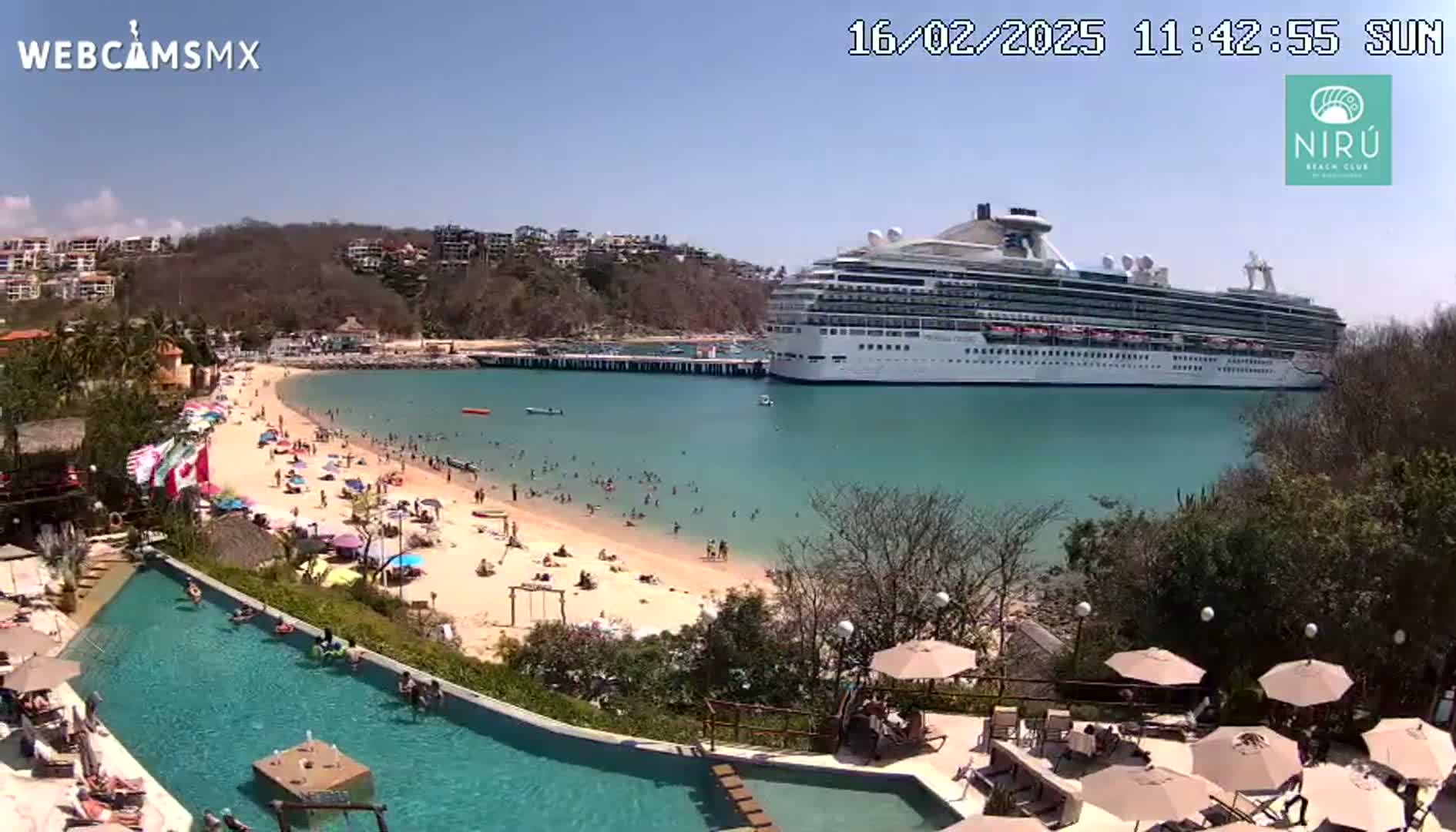 A large cruise ship is docked in a calm, turquoise bay next to a sandy beach with many people swimming and sunbathing; a resort pool and patio are visible in the foreground on a sunny day.
