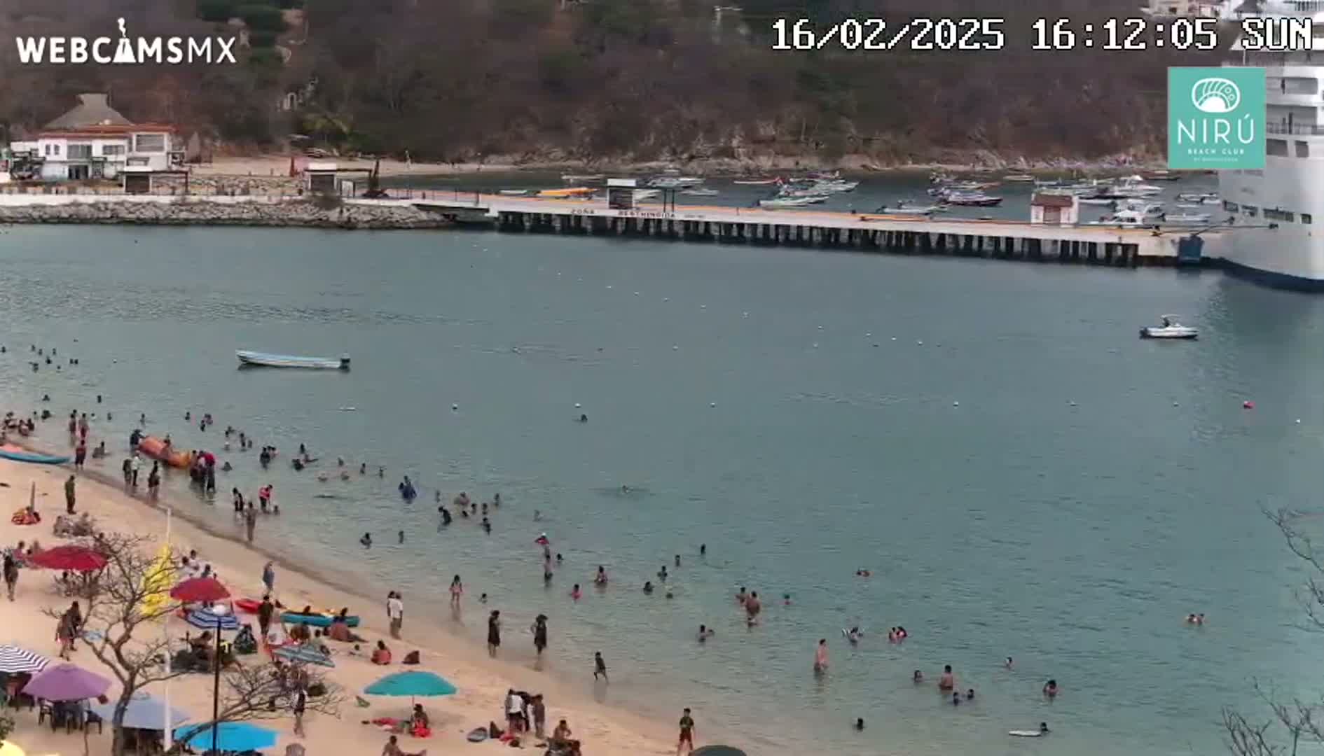 A sandy beach with many people swimming and sunbathing is next to a calm bay with boats and a docked cruise ship, under a sunny sky.