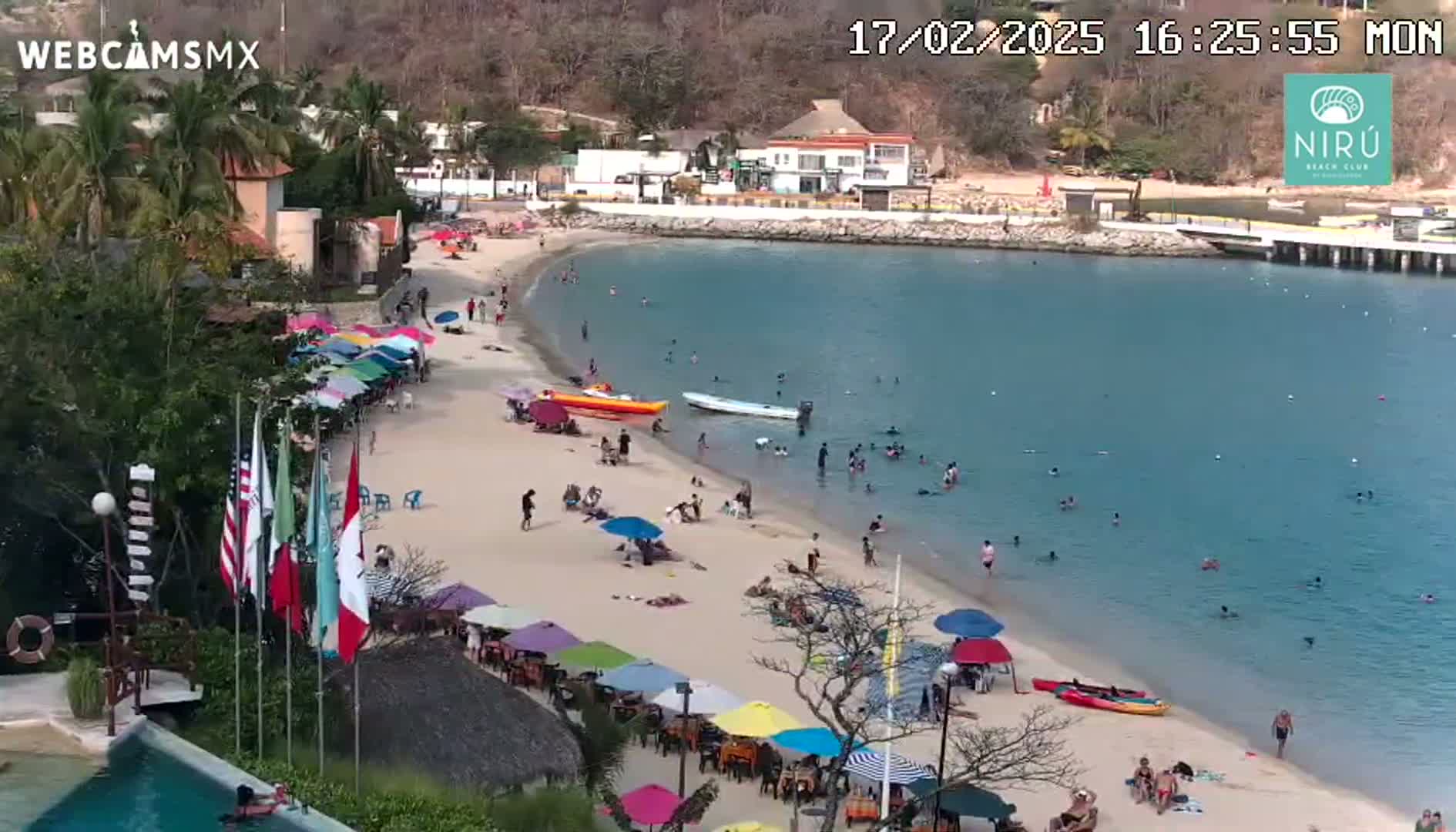 A sandy beach with colorful umbrellas and many people swimming and relaxing in calm, clear water, under a sunny sky.