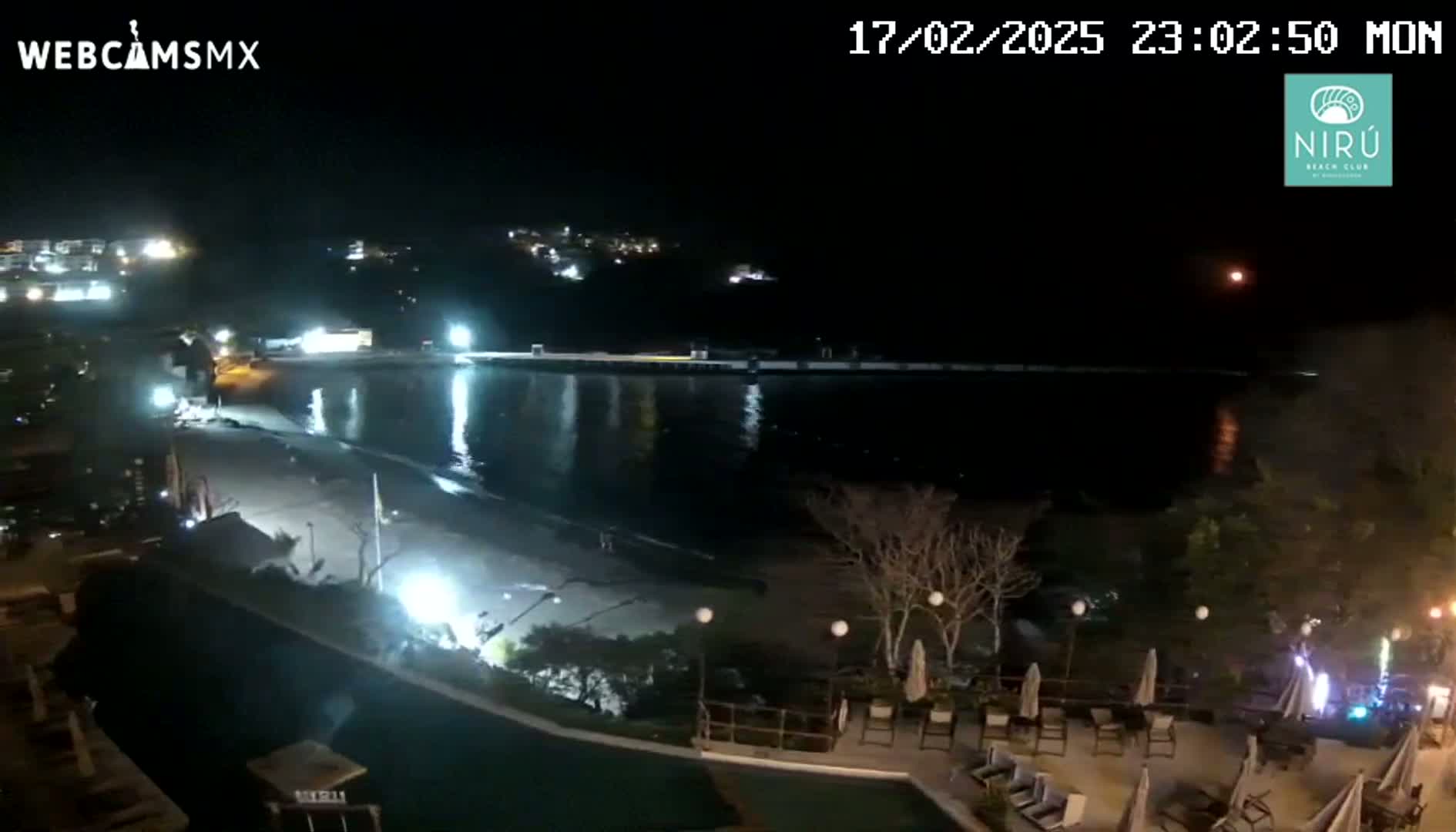 A nighttime view of a beach with a calm body of water, a pier, and a resort area with outdoor furniture under a clear, moonlit sky.