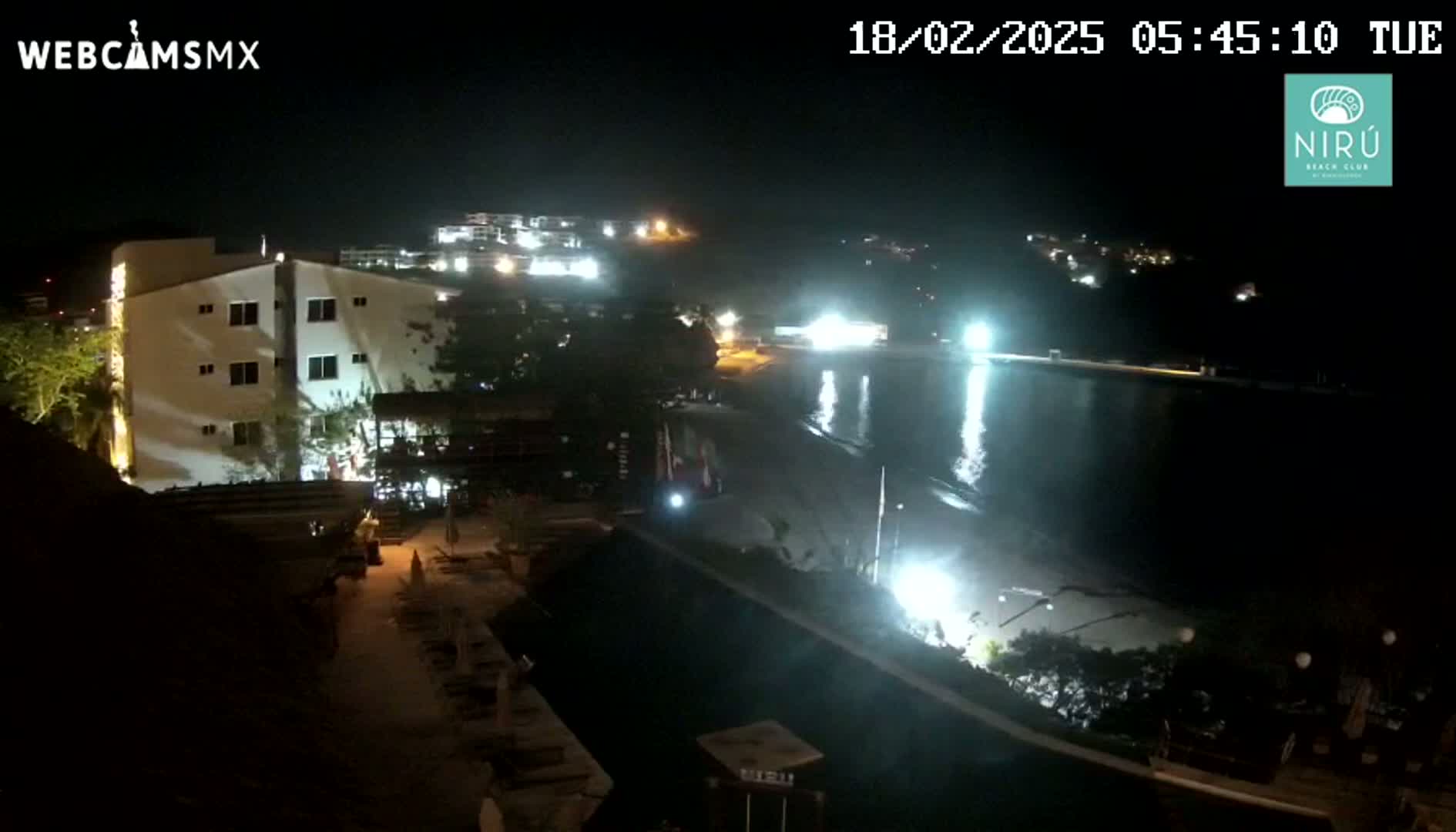 A nighttime view of a beach, lit by artificial lights, with a multi-story building and other structures visible nearby.