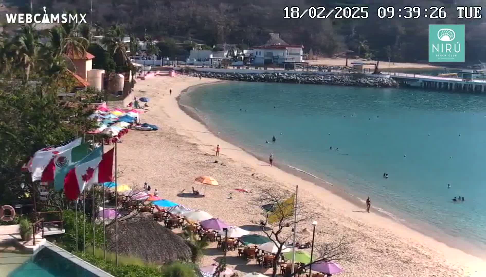 A sandy beach with turquoise water and many colorful umbrellas lines a calm, sunny bay,  with a few people swimming and sunbathing.