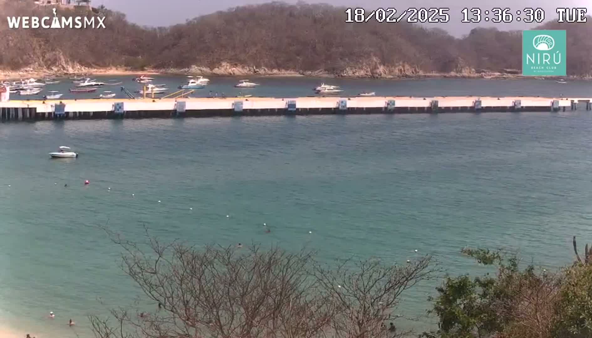 A calm, clear body of water with several boats docked at a pier, surrounded by dry, brown hills under a sunny sky.
