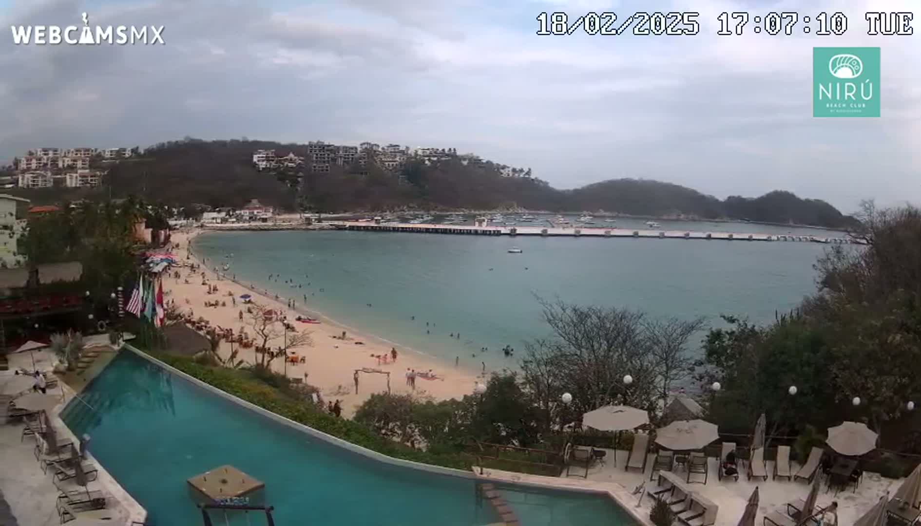 A partly cloudy day shows a sandy beach with swimmers and sunbathers, a calm bay with a pier, a hillside with buildings, and a resort pool area with lounge chairs and umbrellas.