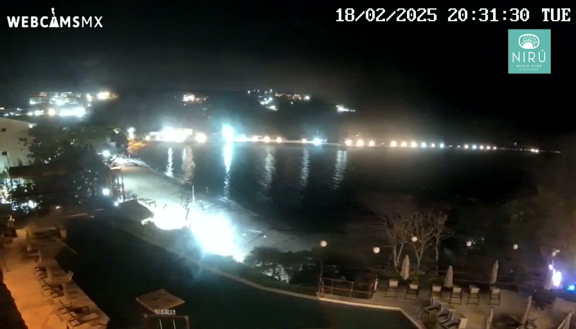 A nighttime view of a beach at a resort with calm water, lights illuminating the shore and buildings in the distance.
