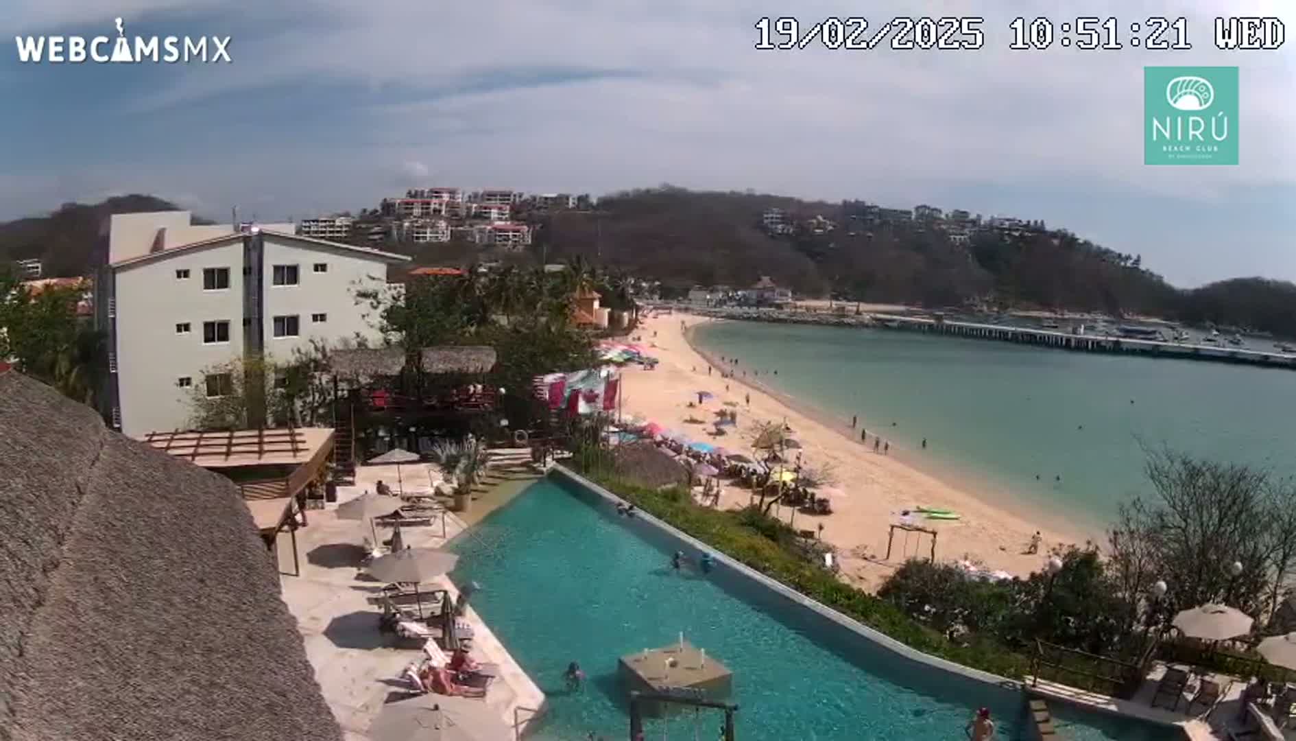A sunny day overlooking a resort pool and a sandy beach with calm, clear water and people swimming and relaxing.