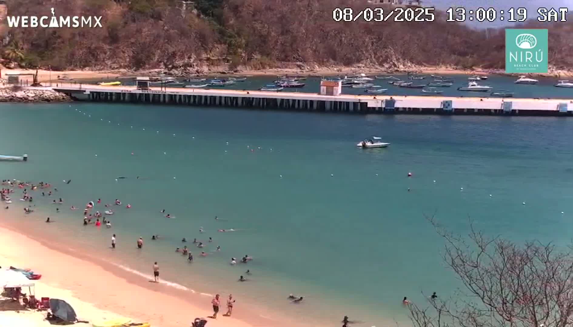 A sandy beach with many people swimming and sunbathing is situated next to a calm, teal ocean with a long pier and several boats moored nearby, under a sunny sky.