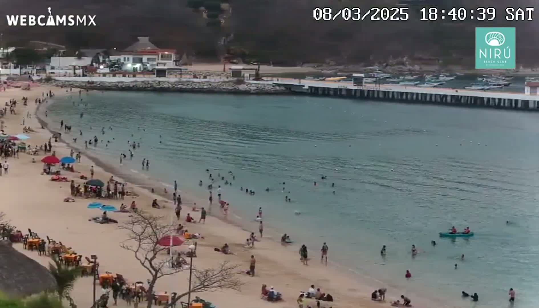 A sandy beach with many people swimming and relaxing,  backed by buildings and a pier, on a calm, overcast day.