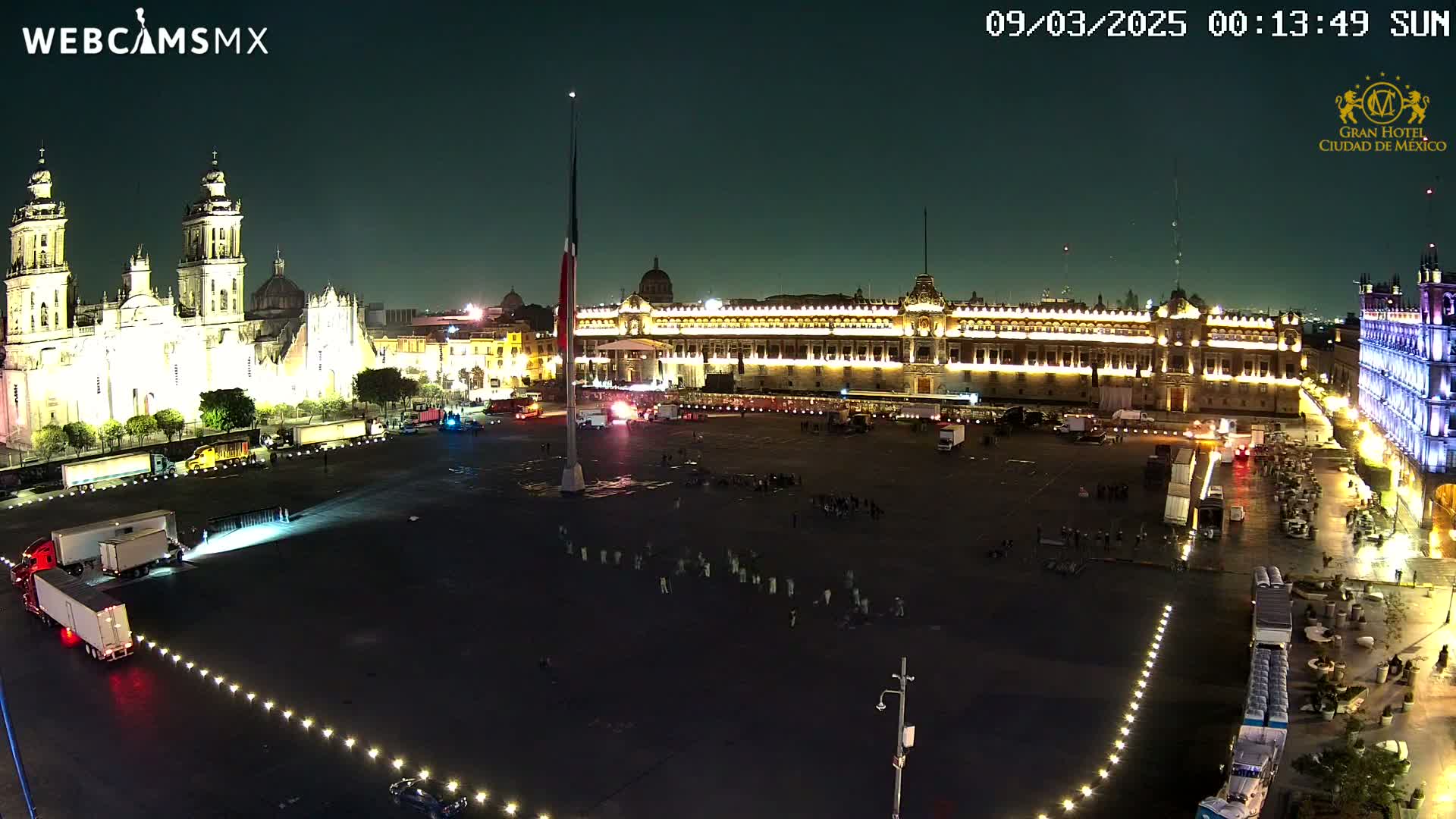 A large, illuminated plaza at night, featuring a cathedral and government buildings, with several trucks and a few people present.