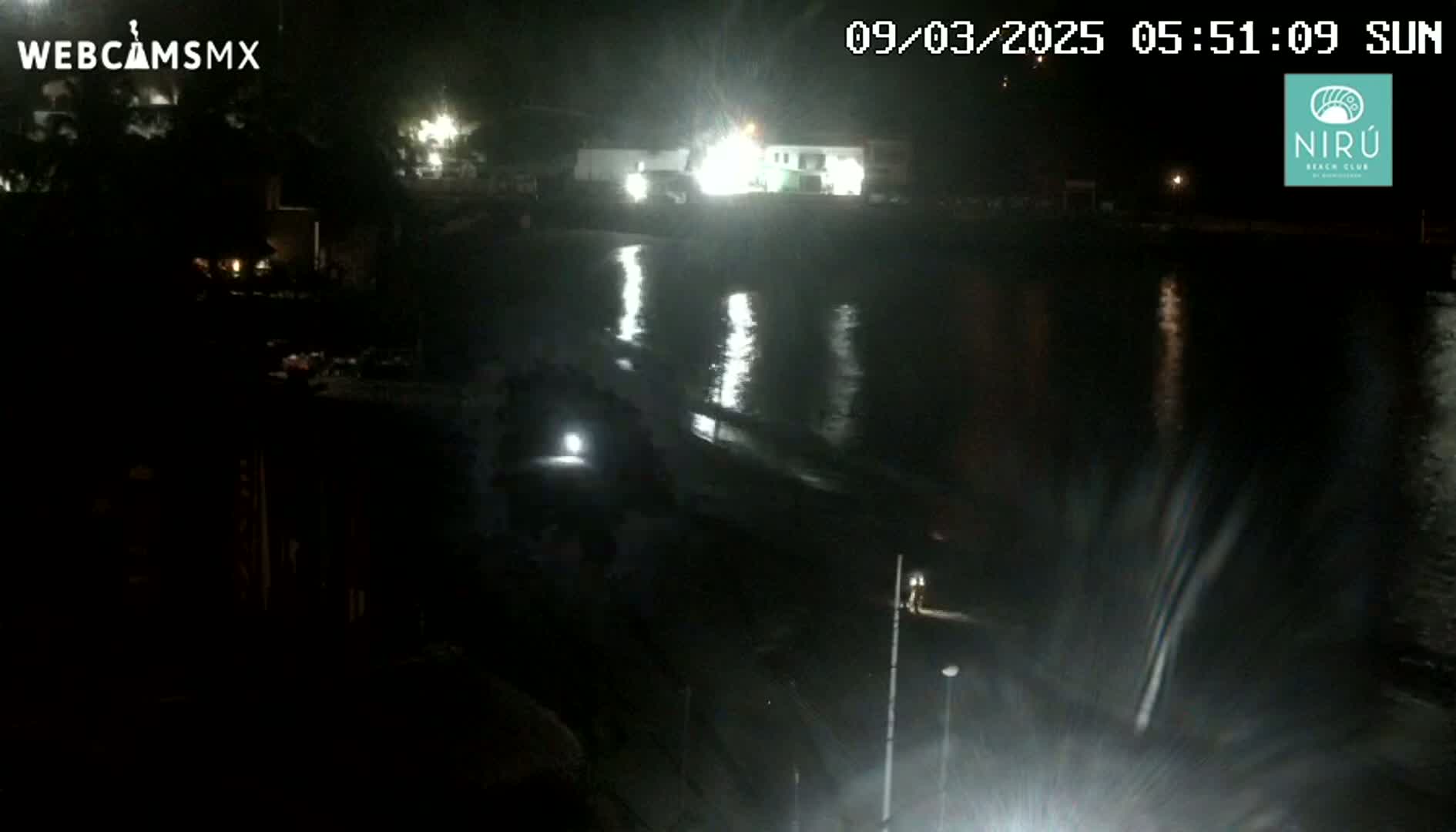 A nighttime view of a dark, calm body of water reflecting lights from buildings and vehicles on the shore.