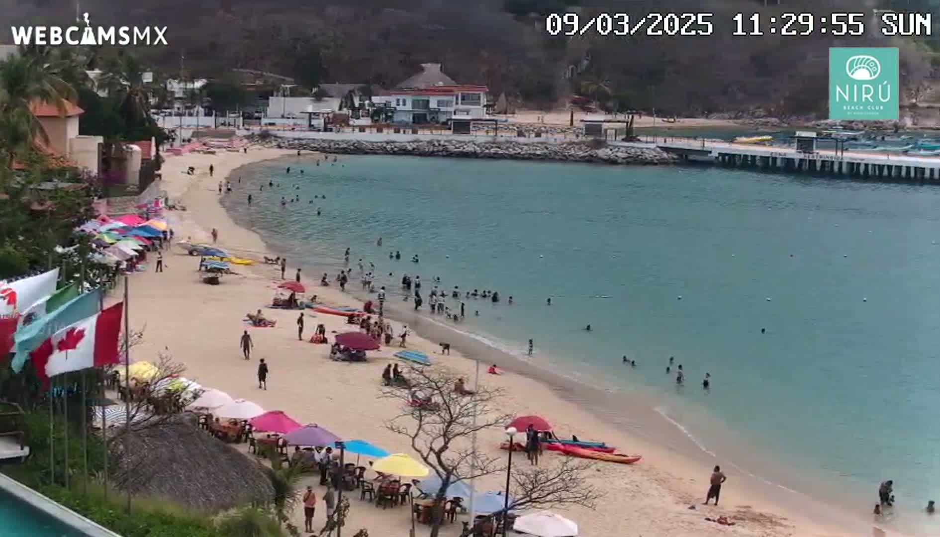A sandy beach with numerous people swimming and relaxing under colorful umbrellas, adjacent to calm, clear water and a small town on a sunny day.
