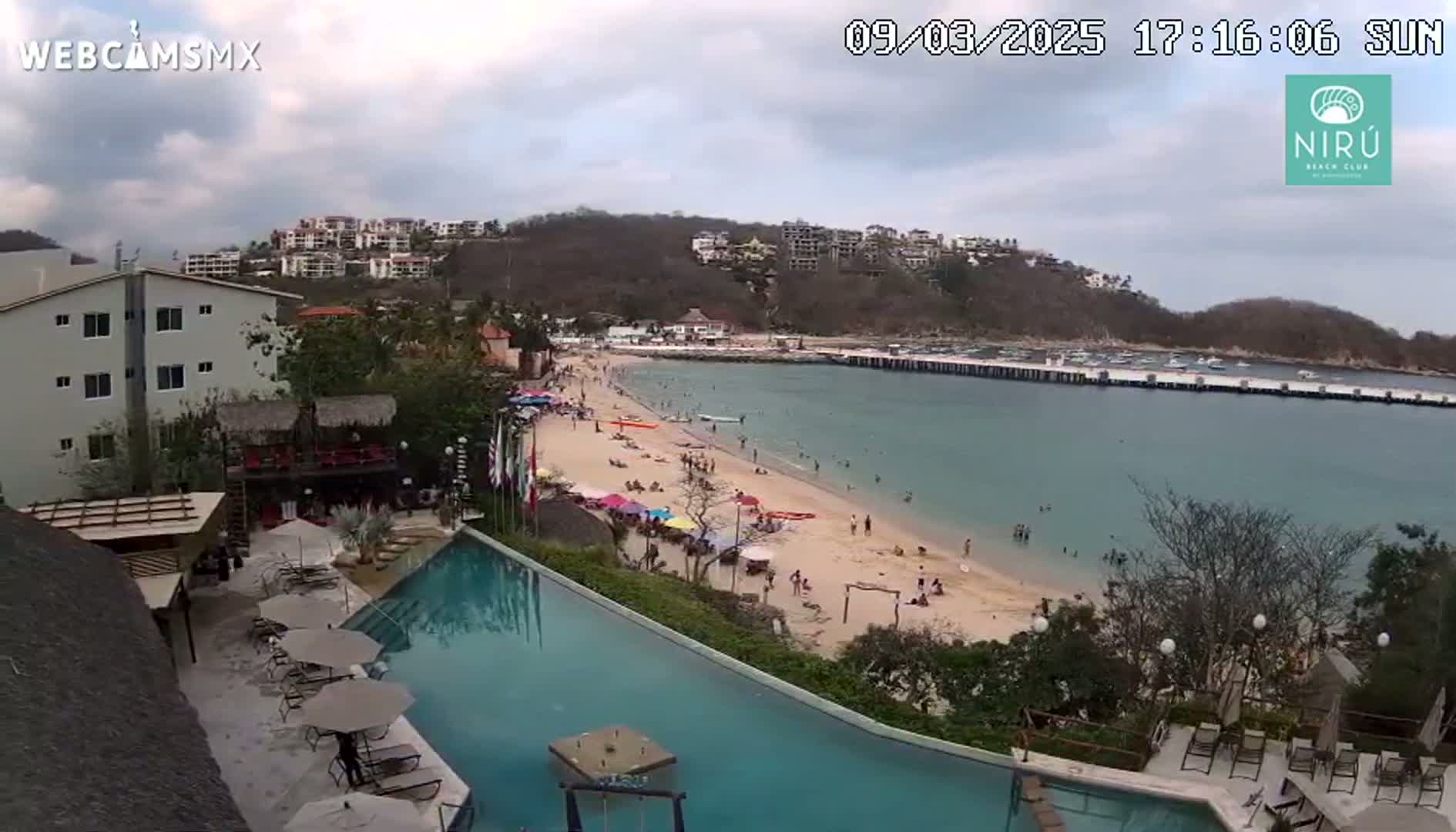 An aerial view of a resort with a long, infinity-style pool overlooking a sandy beach and calm ocean under a partly cloudy sky.