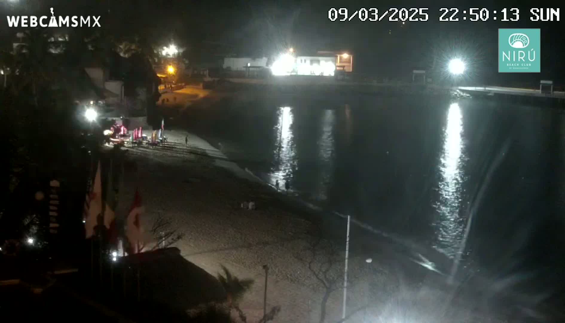 A nighttime view of a mostly empty beach with calm water reflecting lights, under clear skies.