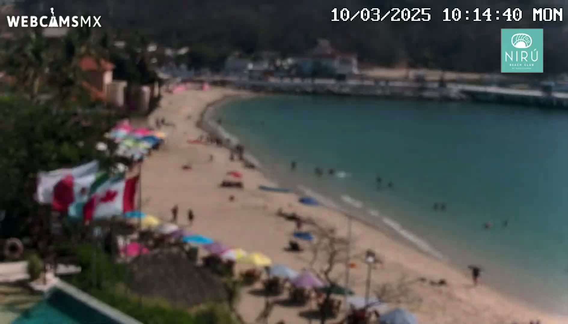 A sandy beach with colorful umbrellas and people, next to calm, teal-colored water, under a sunny sky.