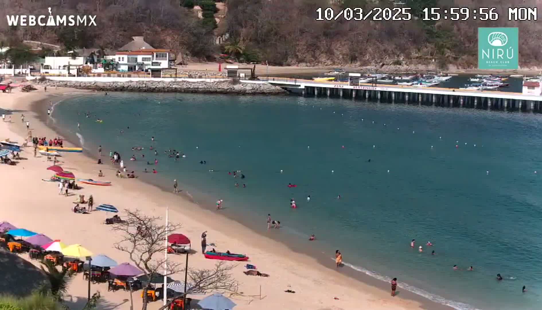 A sandy beach with many people swimming and relaxing under umbrellas, next to a calm, clear blue bay with a pier and several boats.