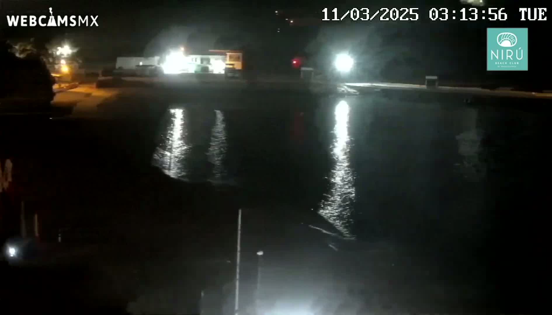 At night, a dark body of water reflects lights from buildings and structures on the shore.