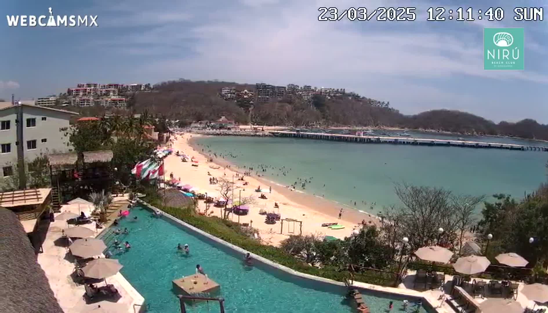 An aerial view shows a resort pool overlooking a sunny beach with many people swimming and relaxing on a clear day.
