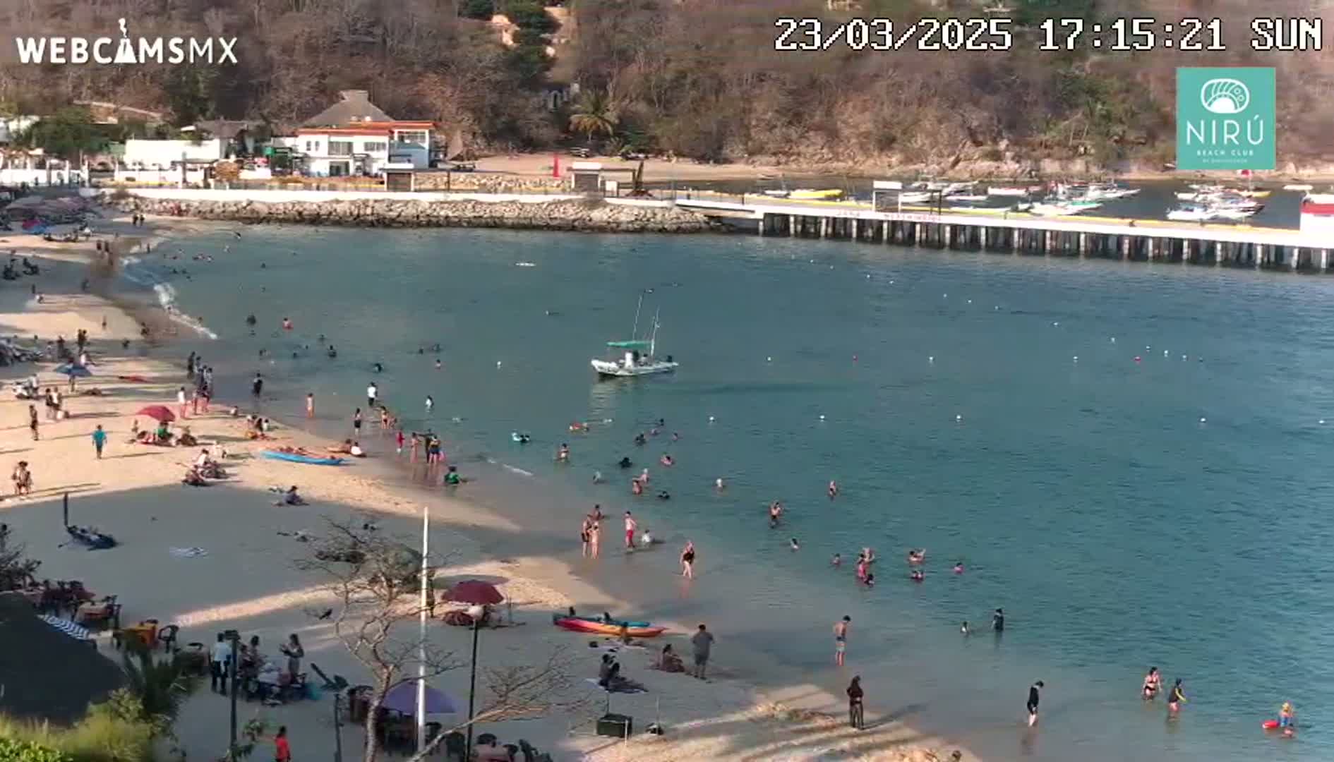 A sandy beach with many people swimming and sunbathing, next to a calm bay with a small boat and a pier, on a sunny day.