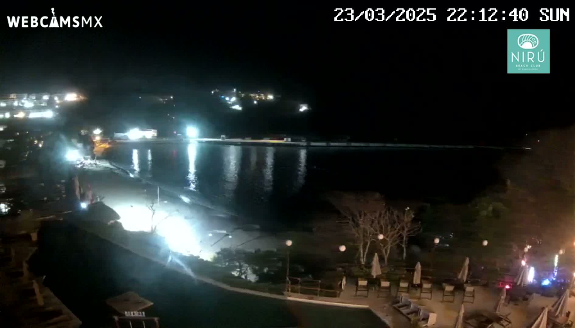 A nighttime view of a beach at a resort with calm water, lights illuminating the sand, and sparsely lit landscaping surrounding a pool area.