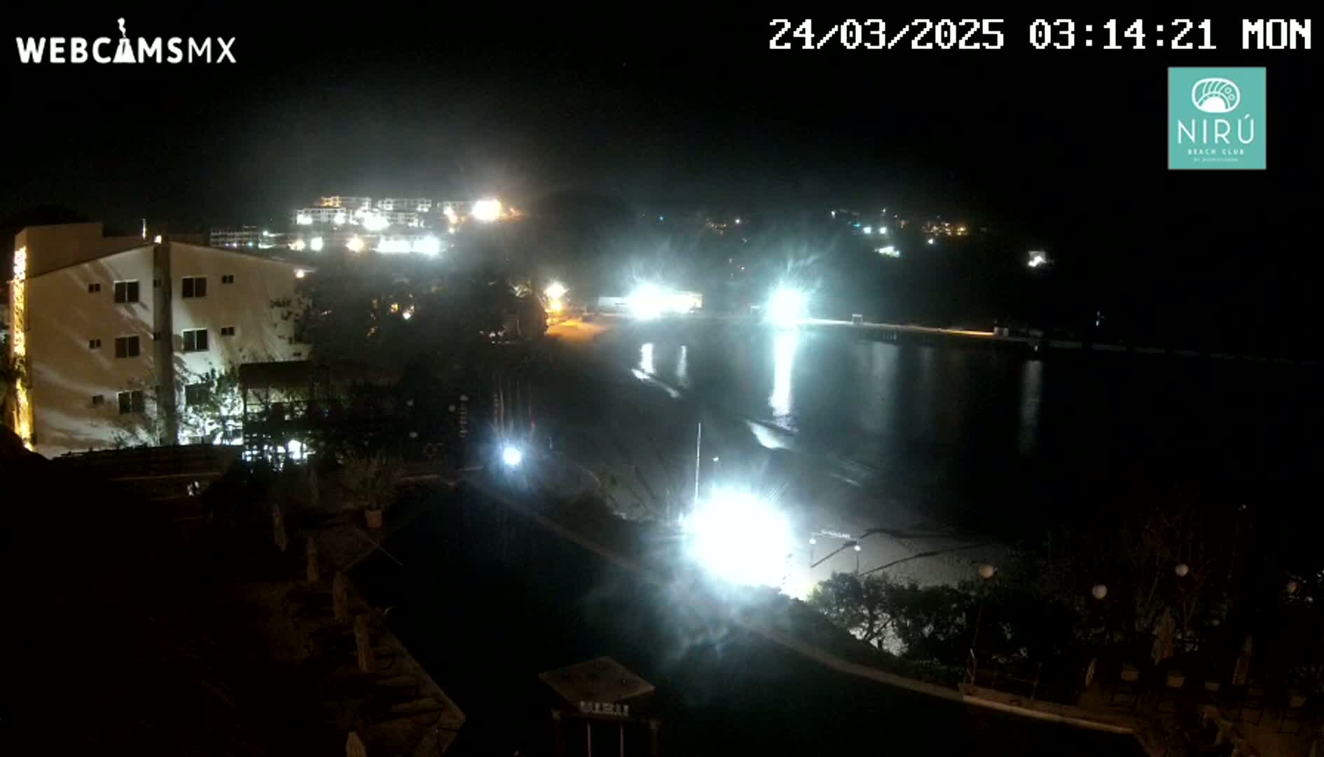 A nighttime view of a beach at a resort, showing brightly lit buildings and a dark ocean reflecting lights.