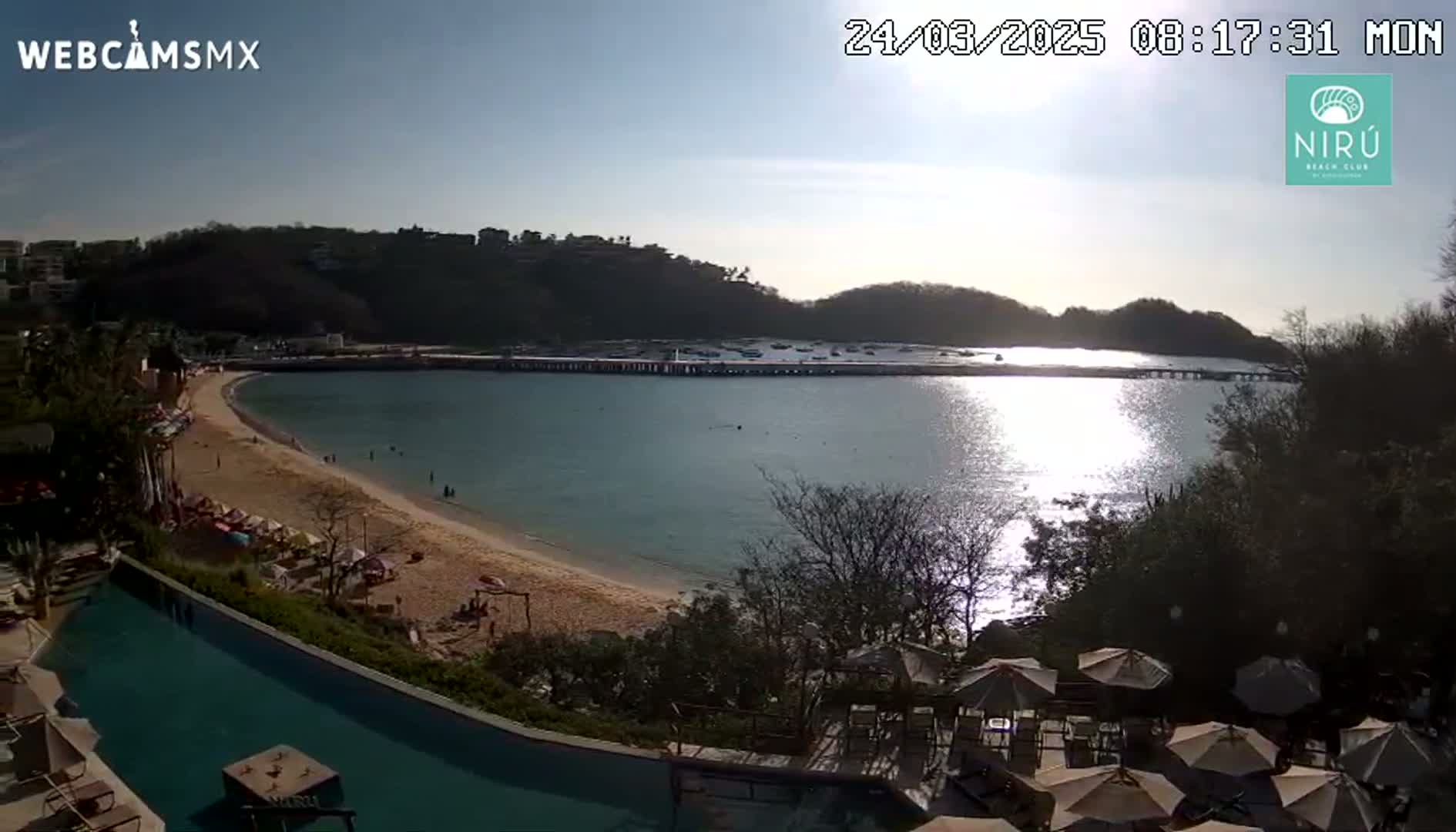 A sunny day at a beach with calm water, a sandy shore, a long pier, and some buildings on a hill overlooking the ocean, seen from an elevated position showing a swimming pool in the foreground.