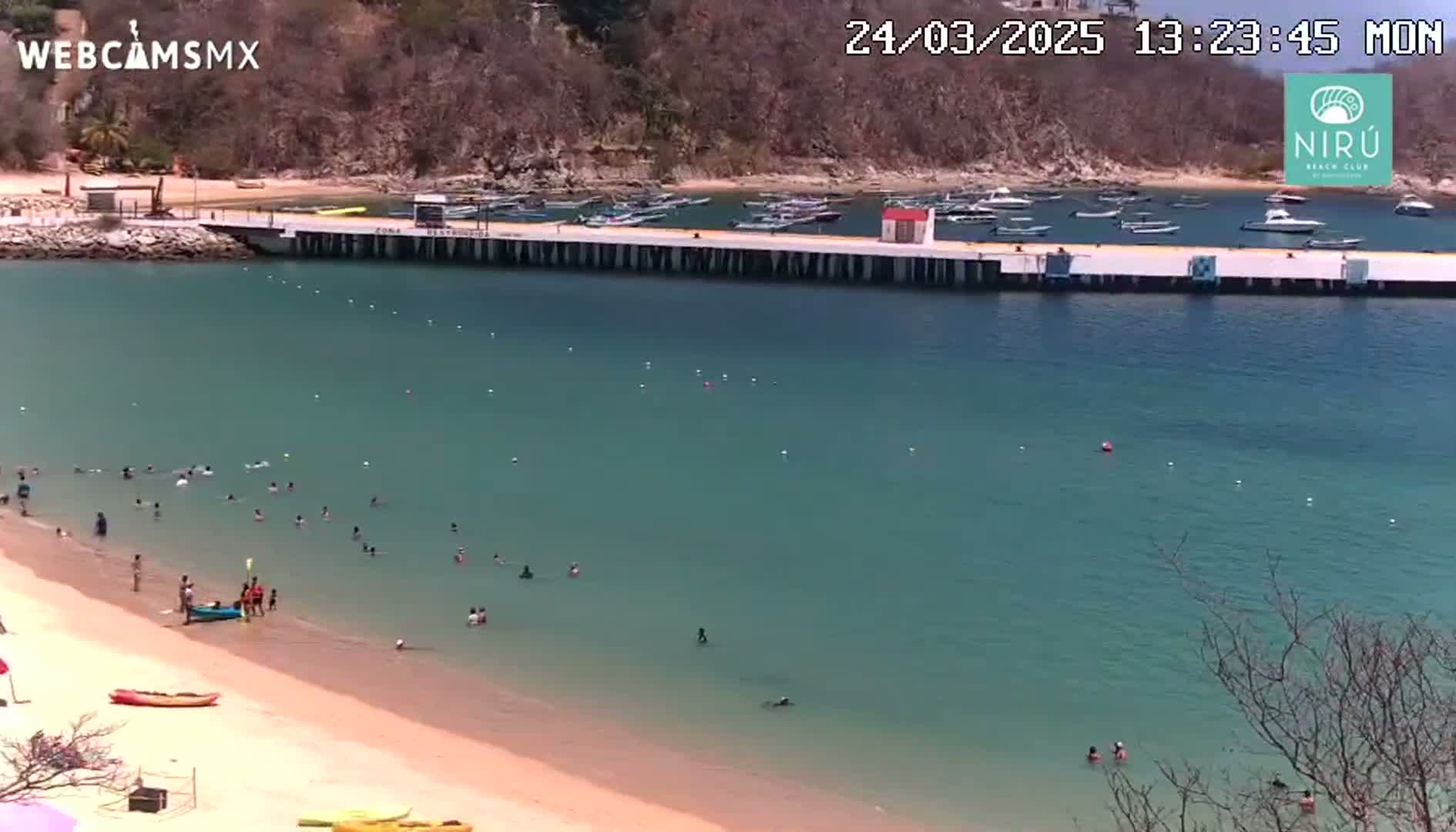 A sandy beach with calm, clear teal water, many people swimming and sunbathing, boats moored at a long pier, and hills in the background under sunny skies.