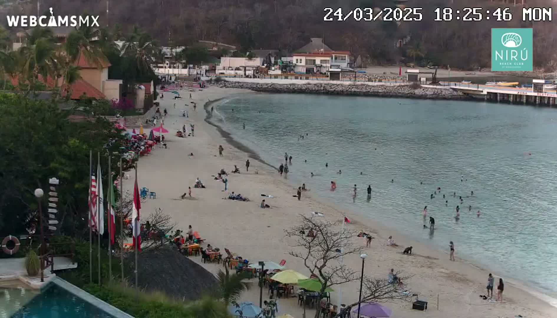 A sandy beach with numerous people swimming and relaxing, situated near buildings and calm water under a partly cloudy sky.