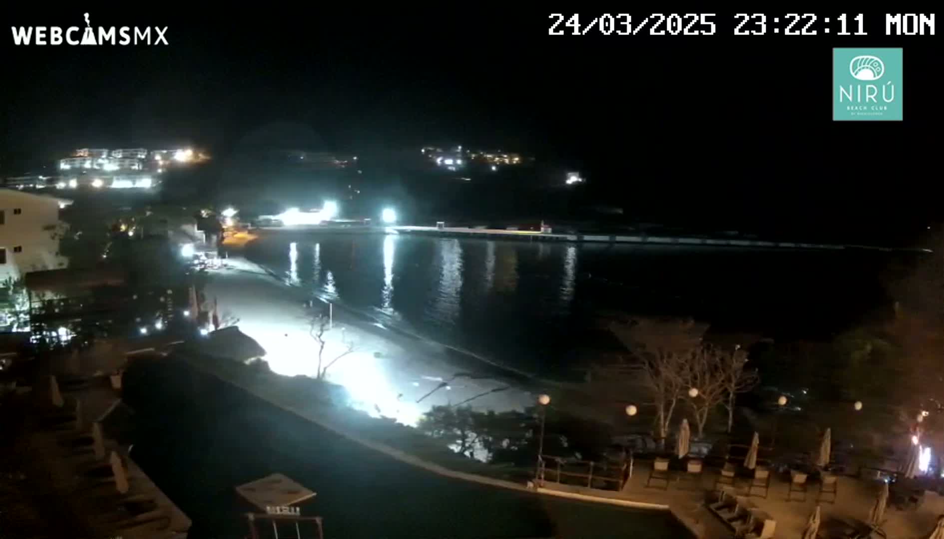 A nighttime view of a beach and waterfront area with buildings and lights illuminating the scene under a clear, dark sky.
