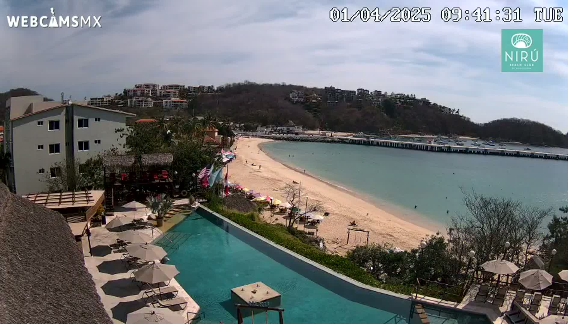 An aerial view of a resort with a swimming pool overlooking a sandy beach and calm ocean under a partly cloudy sky.