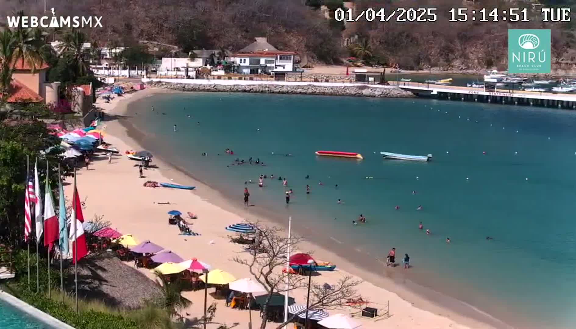 A sunny beach scene shows a sandy shore with colorful umbrellas and people swimming and relaxing in clear turquoise water, with buildings and hills visible in the background.