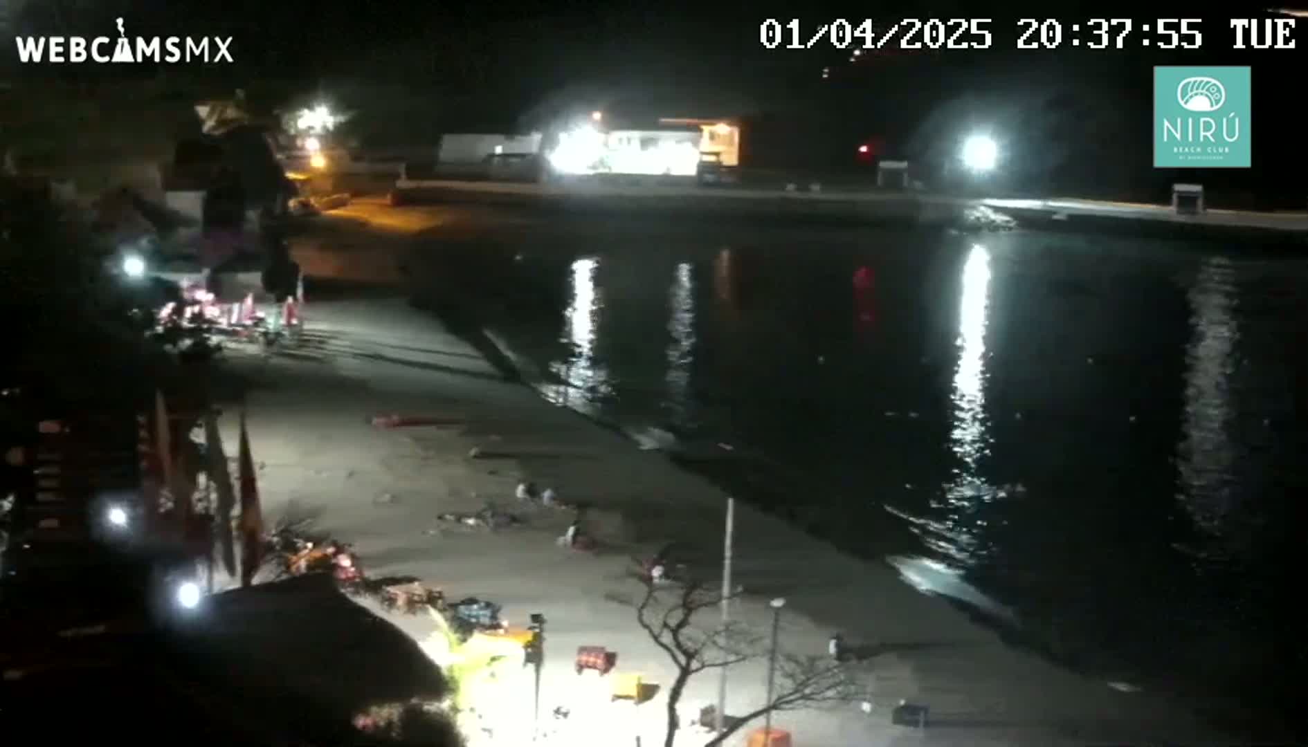 A nighttime, high-angle view shows a mostly empty beach with a few people scattered along the shoreline, next to a calm body of water reflecting bright lights.