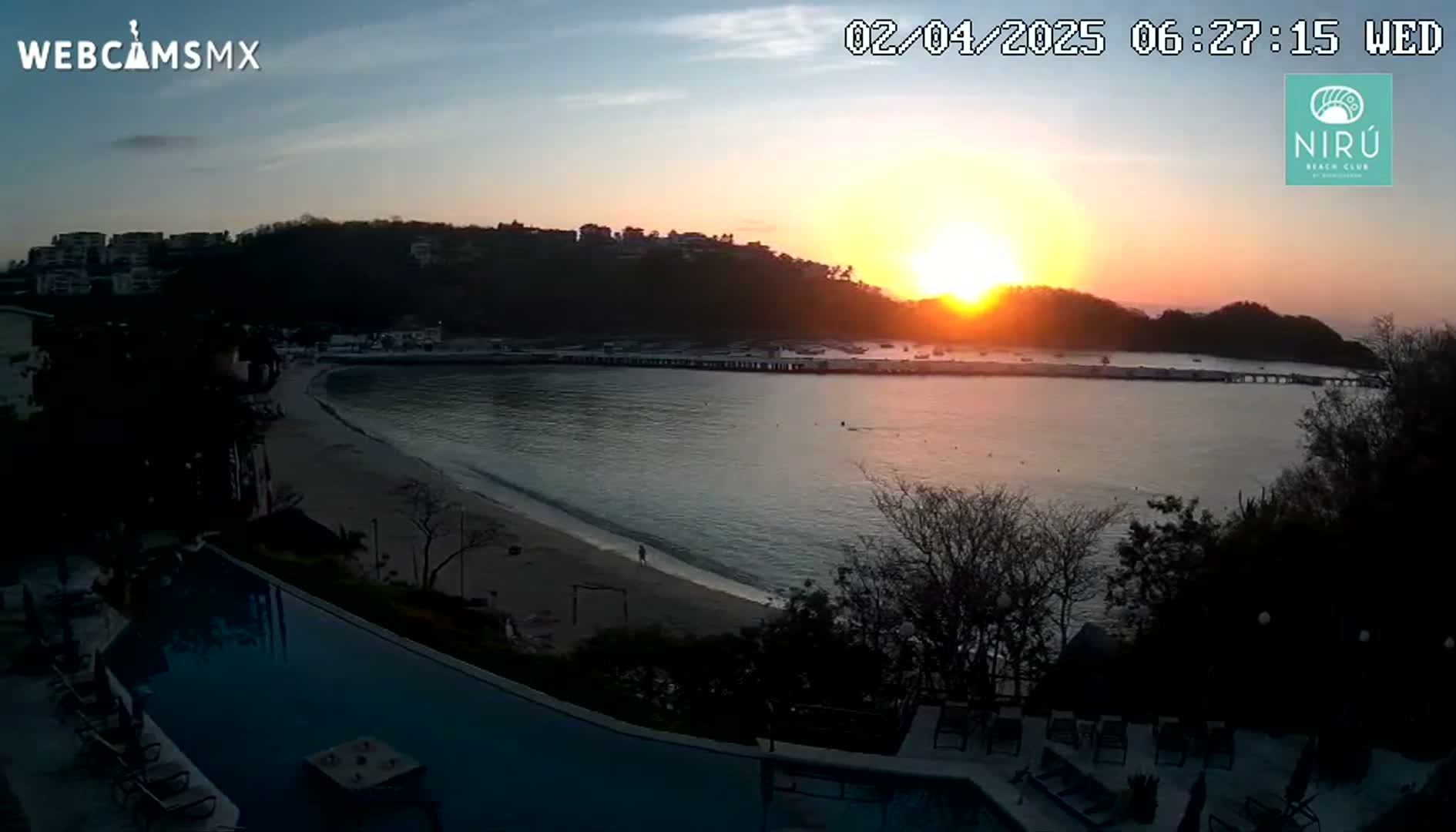 A sunrise over a calm bay with a sandy beach, seen from an elevated viewpoint overlooking a swimming pool and buildings.
