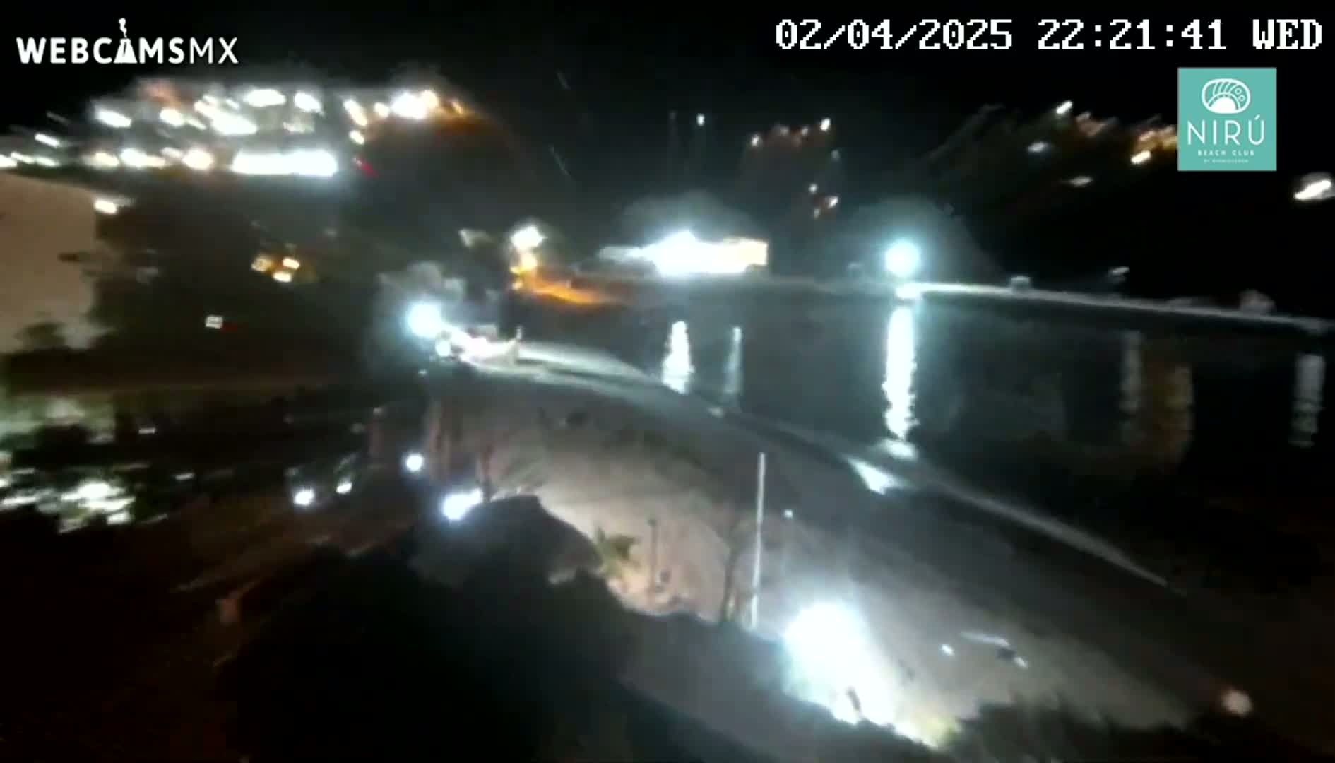 A nighttime, slightly blurry, low-angle view shows brightly lit buildings and vehicles near a body of water with some ripples.
